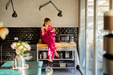 Pembe elbiseli bir kadın mutfak tezgahında oturup telefonuyla sessiz bir sabahın tadını çıkarıyor. Güneş ışığı cam kapılardan geçer, sıcak ve huzurlu bir atmosfer yaratır.