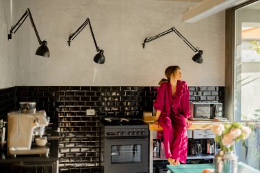 Pembe elbiseli bir kadın mutfak tezgahına çıplak ayakla oturur ve sabah güneşinin tadını çıkarır. Modern siyah kiremitli mutfak ve yumuşak doğal ışık sıcak ve davetkar bir ortam yaratır.