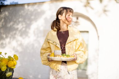 Beyaz bir trullo 'nun önünde kahvaltı için sofrayı hazırlayan kadın. Limonlu ve çiçekli taş bir masa huzurlu Akdeniz sabah manzarasına cazibe katar.