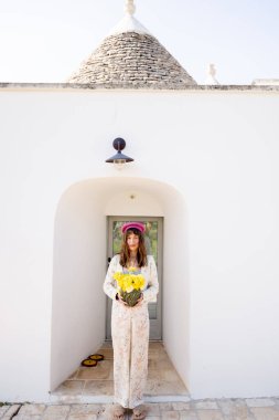 Çiçekçi kıyafeti ve pembe bereli bir kadın güney İtalya 'da güneşli bir bahar gününde elinde parlak sarı ranunculus çiçekleri olan beyaz bir trullo evinin girişinde duruyor.