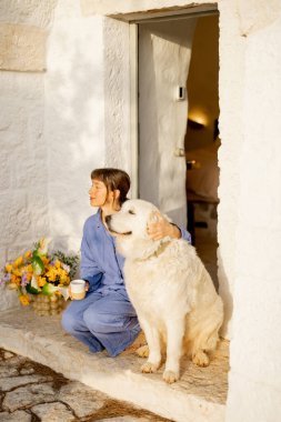 Mavi keten giymiş bir kadın ve sadık beyaz köpeği arasında paylaşılan huzurlu bir an. İtalya 'nın Puglia kentindeki geleneksel bir trullo evinin kapısında huzurlu bir gündoğumu.