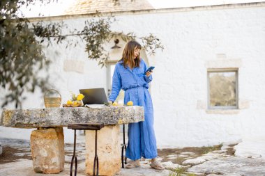 Mavi elbiseli bir kadın dışarıda, taşra bir masada dizüstü bilgisayarın yanında dikilirken görüntülü görüşme sırasında el sallıyor. Resim, yavaş yaşam ve bağlantı dokunuşuyla uzaktan çalışmayı gösteriyor