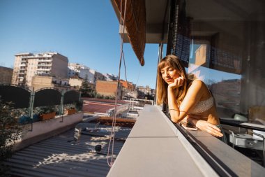 Modern bir apartmanın penceresinden sarkan bir kadın, Roma 'da sabah güneşinin tadını çıkarıyor. Işıkla ve kentsel sakinlikle dolu sessiz, yansıtıcı bir an.