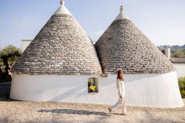 Çiçek elbiseli ve pembe bereli bir kadın Puglia, İtalya 'da sabah güneşinde yıkanmış, konik taştan çatıları olan geleneksel bir beyaz trullo' nun önünden geçiyor.