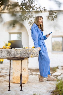 Mavi elbiseli bir kadın dışarıda, taşra bir masada dizüstü bilgisayarın yanında dikilirken görüntülü görüşme sırasında el sallıyor. Resim, yavaş yaşam ve bağlantı dokunuşuyla uzaktan çalışmayı gösteriyor
