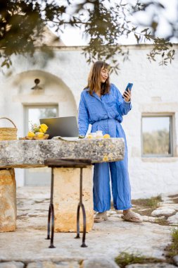 Mavi elbiseli bir kadın dışarıda, taşra bir masada dizüstü bilgisayarın yanında dikilirken görüntülü görüşme sırasında el sallıyor. Resim, yavaş yaşam ve bağlantı dokunuşuyla uzaktan çalışmayı gösteriyor