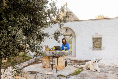 Bir kadın köpeğiyle birlikte trullo evinin önündeki taştan bir masada dizüstü bilgisayarıyla çalışıyor. Konsept uzak çalışmaları, dijital göçebe yaşam tarzını ve barışçıl doğa bağlantısını yakalıyor