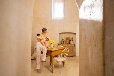 Bir kadın taştan bir mutfaktaki ahşap bir masada sarı çiçekler düzenliyor. Sahne basitlik, gelenek ve konforlu Akdeniz yaşamını iç mekan veya yaşam tarzı kullanımı için mükemmel bir şekilde yansıtıyor