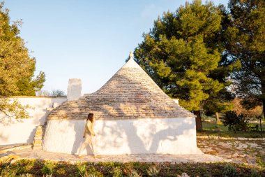 Puglia 'da zeytin ağaçları ve yeşilliklerle çevrili güneşli bir trullo' nun yanında yürüyen bir kadın. Bu resim seyahat refahı veya kırsal yaşam kavramlarında kullanmak için miras ve yaşam tarzını harmanlıyor