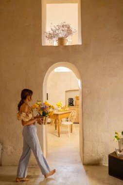 Bir kadın taştan bir mutfaktaki ahşap bir masada sarı çiçekler düzenliyor. Sahne basitlik, gelenek ve konforlu Akdeniz yaşamını iç mekan veya yaşam tarzı kullanımı için mükemmel bir şekilde yansıtıyor