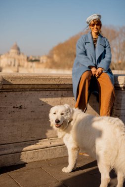 Roma 'da güneşli bir kış yürüyüşü sırasında neşeli bir an. Tiber 'in yanındaki taş bir duvarda, mavi bir paltoyla sarmalanmış, tüylü beyaz köpeğimle altın ışığın tadını çıkarıyorum.