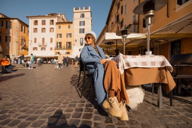 Mavi ceketli gülümseyen kadın, sıcak tonlu binalarla çevrili, güneşli bir Roma meydanında kahvesinin tadını çıkarıyor.