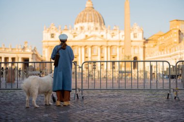 Mavi paltolu bir kadın ve beyaz köpeği Roma 'daki St. Peters Meydanı' nın barikatlarının yanında huzur içinde duruyor. Bu görüntü seyahat sükuneti ve ruhani turizmi sembolik bir ortamda yakalıyor.