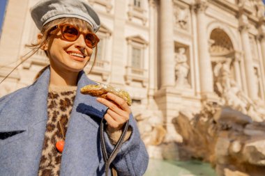 Mavi ceketli neşeli bir kadın Roma 'daki Trevi Çeşmesi' nin önünde geleneksel bir cannolo yiyor. İtalya 'nın lezzet ve güneşini kucaklıyor.