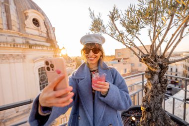 Roma 'nın çatısında elinde kırmızı bir kokteyl tutarak selfie çeken bir kadın. Kubbe ve zeytin ağacı bu canlı şehre cazibe katıyor.