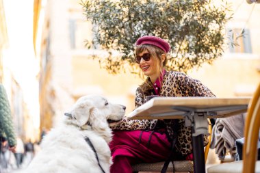 Şık giyinmiş gülümseyen bir kadın beyaz köpeğiyle birlikte Roma 'da bir kafe masasında oturuyor. Görüntü neşe, dostluk ve yavaş şehir yaşamını hayat tarzı ve seyahat içeriği için ideal hale getiriyor.