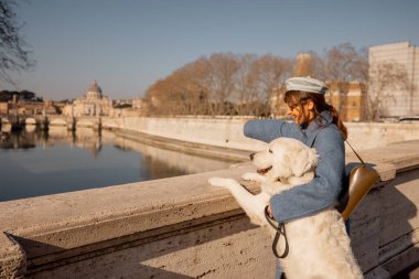 Mavi ceketli bir kadın, büyük beyaz köpeğini bir Roma köprüsünde kucaklıyor. Açık bir kış gökyüzünün altında St. Peters Bazilikası 'na bakıyor.