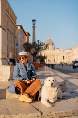 Mavi paltolu ve şapkalı bir kadın köpeğiyle birlikte Saint Peters Bazilikası 'nın yakınlarında güneş ışığının tadını çıkarıyor. Fotoğraf, Roma 'da yavaş seyahat ve evcil hayvan dostu şehir anlarını yakalıyor.