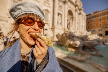 Mavi ceketli neşeli bir kadın Roma 'daki Trevi Çeşmesi' nin önünde geleneksel bir cannolo yiyor. İtalya 'nın lezzet ve güneşini kucaklıyor.