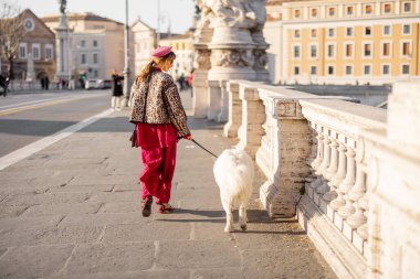 Güneşli bir günde, bir kadın Roma köprüsü boyunca büyük beyaz köpeğini gezdirir. Fotoğraf, yavaş şehir yaşamını gösteriyor ve gezici hayvan yaşam tarzı ve Avrupa şehir terfileri için ideal.