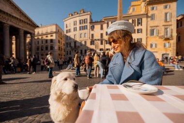 Mavi paltolu bir kadın, Roma meydanındaki bir kafede beyaz köpeğini kucaklıyor. Roma 'nın kalbinde huzurlu bir sevgi anı yakalıyor.