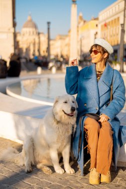 Mavi ceketli gülümseyen bir kadın St. Peter Bazilikası yakınlarındaki büyük beyaz köpeği yanında oturan modern bir çeşmeye bozuk para atıyor. Roma 'da umudun sembolü ve neşeli seyahat anları