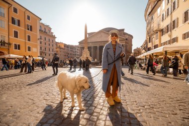 Mavi ceketli şık bir kadın ve tüylü beyaz köpeği Pantheon 'un önünde duruyor, Roma güneşinin altında canlı kalabalıkların ve kaldırım taşlı sokakların arasında güneşleniyor.