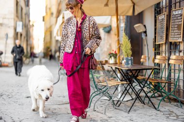 Kalın renkli şık bir kadın tüylü beyaz köpeğiyle büyüleyici bir Roma kafesinin önünden geçiyor. Sahne, sakin bir şehir gezisi ve İtalya 'daki günlük hayatın zarafetini yakalıyor.