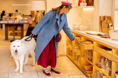 Pembe bereli ve mavi ceketli bir kadın köpeğini şık raflarla çevrili modern bir butikte gezdirir. Moda, evcil hayvanlar ve lüks perakende için ideal