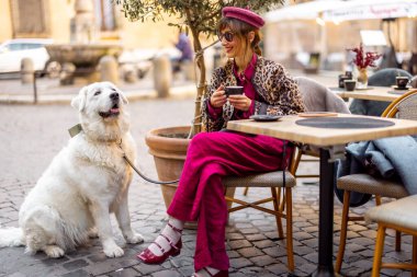 Canlı giysiler içinde şık bir kadın Roma 'da açık bir kafede beyaz köpeğiyle oturuyor. Görüntü yavaş yaşam tarzını ve Avrupa kentsel hayvan dostu kültürünü yansıtıyor