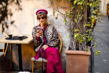 Leopar desenli ceketli ve pembe elbiseli şık bir kadın Roma kafesinin önünde telefonuna bakıyor. Bu görüntü modern şehir yaşam tarzını ve sokak modasını yansıtıyor.