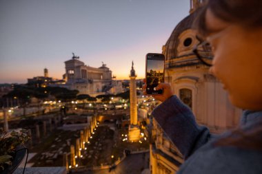 Mavi ceketli kadın günbatımında Roma siluetini yakalıyor. Arka planda Trajans Sütunu ve Vittoriano var. Manzaralı bir çatıda sıcak bir akşam ışığında parlıyor.