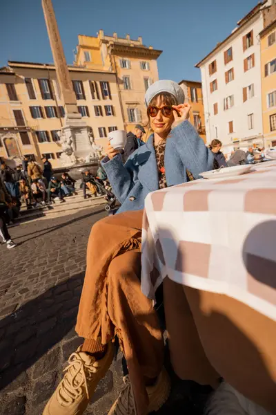 Mavi ceketli bir kadın sabah kahvesini Roma 'daki Pantheon' un önündeki masada içiyor. Güneşin ve hayat dolu sokak hayatının tadını çıkarıyor.