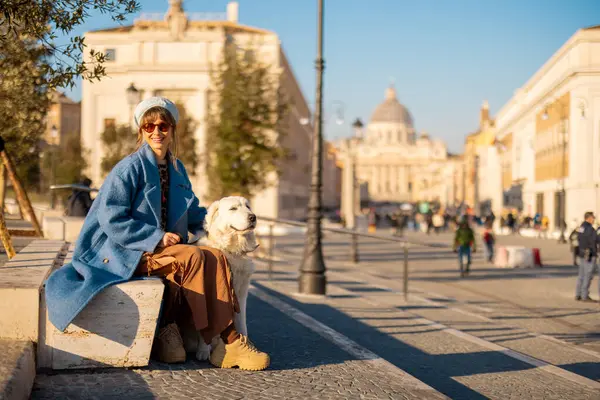Mavi paltolu kadın, büyük beyaz köpeği yanında kestirirken Roma caddesinde oturuyor. Bu resim rahatlama, arkadaşlık ve şık şehir turizmini betimliyor