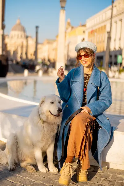 Mavi ceketli gülümseyen bir kadın St. Peter Bazilikası yakınlarındaki büyük beyaz köpeği yanında oturan modern bir çeşmeye bozuk para atıyor. Roma 'da umudun sembolü ve neşeli seyahat anları