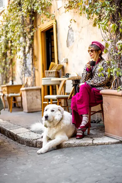Klasik kıyafetli bir kadın büyük beyaz köpeğiyle Roma 'da büyüleyici bir kafede oturuyor. Görüntü, rahat şehir yaşam tarzını seyahat için mükemmel yansıtıyor.
