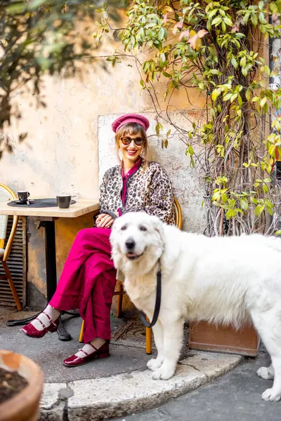 Klasik kıyafetli bir kadın büyük beyaz köpeğiyle Roma 'da büyüleyici bir kafede oturuyor. Görüntü, rahat kentsel yaşam tarzını seyahat ve editör kullanımı için mükemmel yansıtıyor