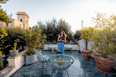 Akdeniz 'deki terasta gün batımının tadını çıkaran bir kadın. Etrafı yeşilliklerle çevrili. Günlük kıyafet, neşeli ifade ve rahat bir yaşam ortamı.