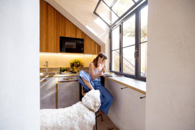 Bir kadın güneşli bir İtalyan mutfağında büyük beyaz köpeğiyle huzurlu bir kahvaltı anını paylaşıyor. Evcil hayvan ürünleri markaları, ev yaşam tarzı veya mutfak tasarımı içeriği için harika
