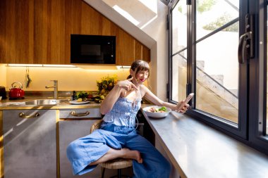 Bir kadın, güneşli bir mutfakta sağlıklı bir kahvaltı yaparken video görüşmesinde el sallıyor. Uzak iş, yaşam tarzı teknolojisi ya da modern yaşam hakkında reklamlar için ideal.