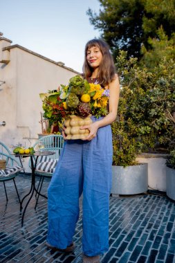 Gülümseyen kadın kiremitli terasta çıplak ayakla yürüyor enginarlar ve limonlarla dolu büyük bir buket tutuyor yeşil ve kırsal cazibeyle çevrili huzurlu güneşli bir günün tadını çıkarıyor.
