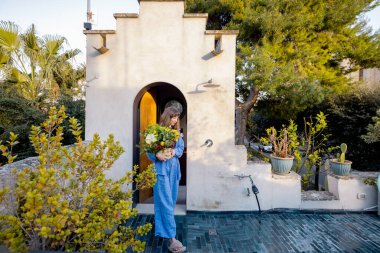 Elinde canlı bir buket tutan genç bir kadın yemyeşil bitkiler, kır taşı ve Akdeniz cazibesiyle çevrili güneşli bir terasa adım atıyor.