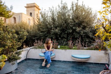 Bir kadın, bahçenin çatısındaki terasta, bitki ve tarihi Akdeniz mimarisi ile çevrili, huzurlu ve yavaş bir yaşam tarzını çağrıştıran, sessiz bir anın tadını çıkarır.