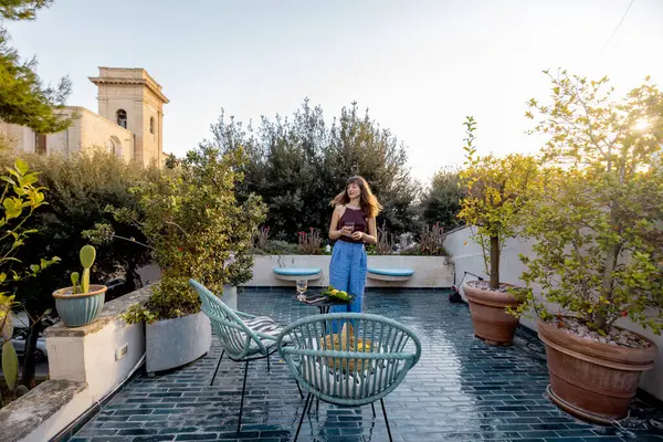 Akdeniz 'deki terasta gün batımının tadını çıkaran bir kadın. Etrafı yeşilliklerle çevrili. Günlük kıyafet, neşeli ifade ve rahat bir yaşam ortamı.