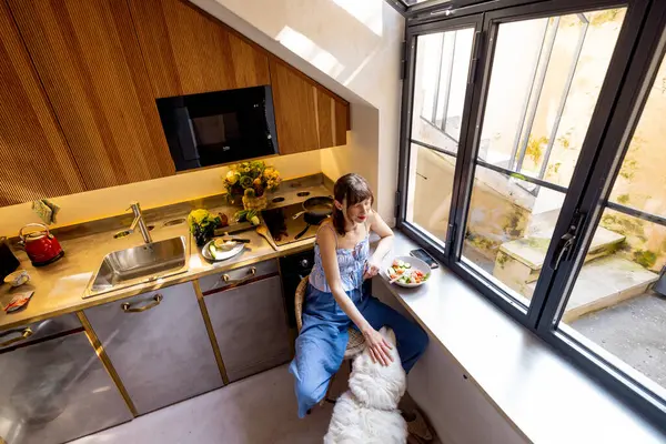 Bir kadın güneşli bir mutfakta köpeğiyle taze bir salatanın ve sessiz bir anın tadını çıkarır. Sağlıklı yaşam, ev hayatı, evcil hayvan dostu yaşam ya da sabah rutin içeriği için harika.