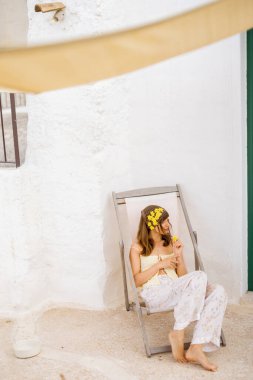 Saçı sarı çiçeklerle dolu sakin bir kadın güneşli beyaz bir duvara yaslanmış şezlongda yalın ayak dinleniyor ve sakin bir Akdeniz öğleden sonrası sıcaklığını kucaklıyor.