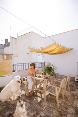 Güney İtalya 'da kırsal çatı terasında taze meyve ve kahveyle yavaş yavaş kahvaltı yapan bir kadın. Yaz ışığı, kaktüs bitkileri ve tüylü köpek dingin sabah havasını tamamlıyor.