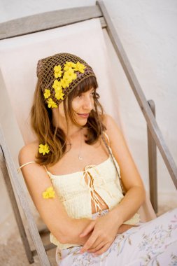 Saçı sarı çiçeklerle dolu sakin bir kadın güneşli beyaz bir duvara yaslanmış şezlongda yalın ayak dinleniyor ve sakin bir Akdeniz öğleden sonrası sıcaklığını kucaklıyor.