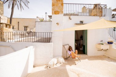 Güneşli bir çatıda bir kitapla rahatlayan bir kadın, kumaş bir gölgeyle gölgelenmiş, yanında büyük beyaz bir köpek, büyüleyici bir güney İtalya kasabasında sakin bir yaz anı.