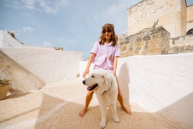 Genç bir kadın ve tüylü beyaz köpeği tarihi taş duvarlar ve saksı bitkileriyle çevrili bir Akdeniz çatısı üzerinde duruyorlar. Güney İtalya 'da sakin ve güneşli bir an